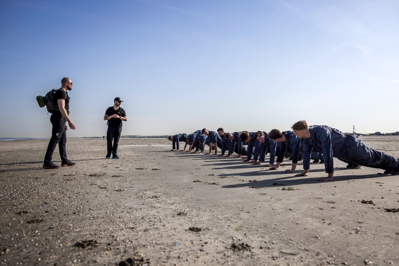 Twee instructeurs kijken toe terwijl een lange rij deelnemers zich opdrukt op het strand